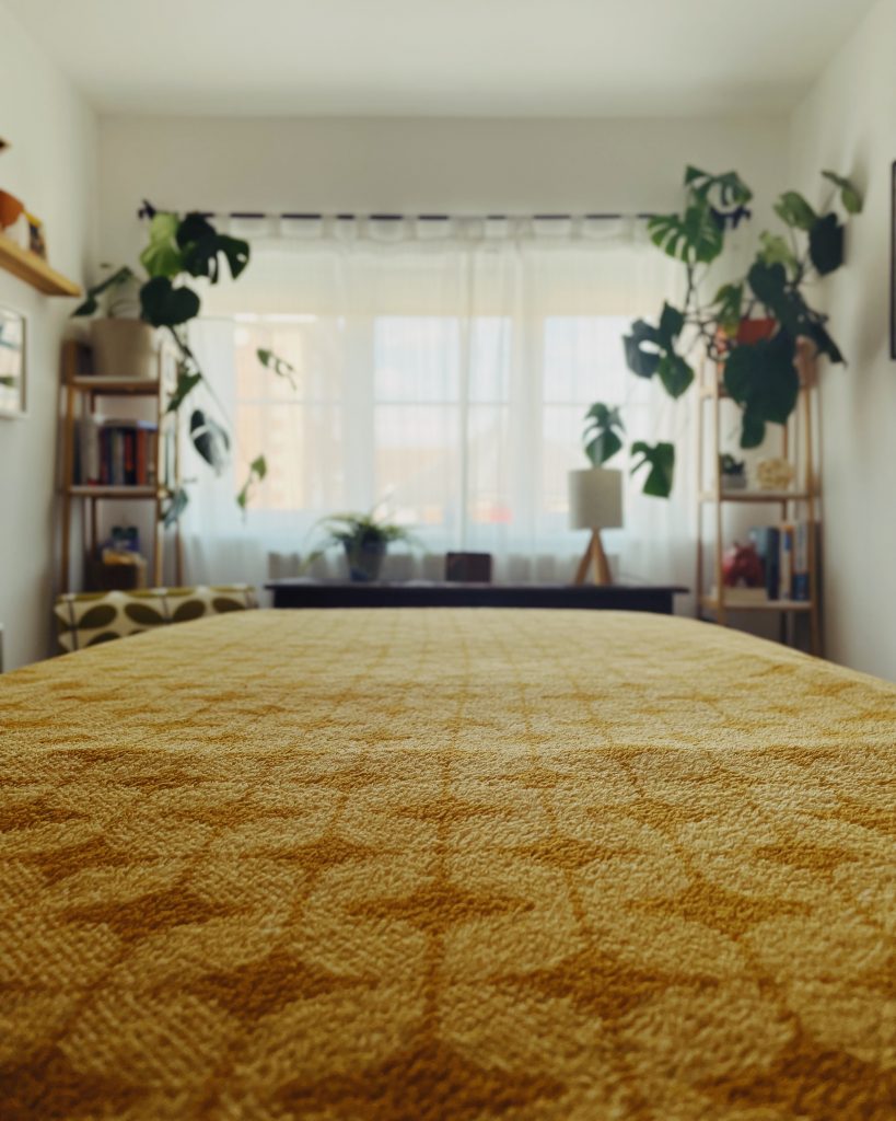View from the bottom of a therapy couch covered in a yellow golden blanket with geometric shapes. At the end of the bed is a window covered with sheet white curtains and either side are plants on shelves.