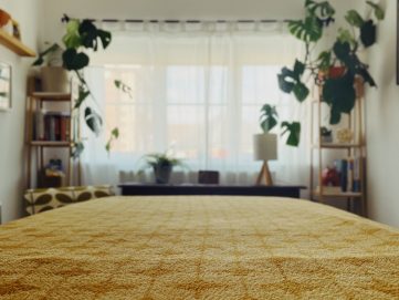 View from the bottom of a therapy couch covered in a yellow golden blanket with geometric shapes. At the end of the bed is a window covered with sheet white curtains and either side are plants on shelves.