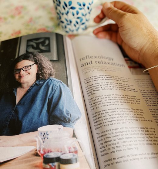 A hand resting on an open magazine with a cup of tea. The magazine is open on an article featuring an image of Sarah Woodhouse with the text about Reflexology