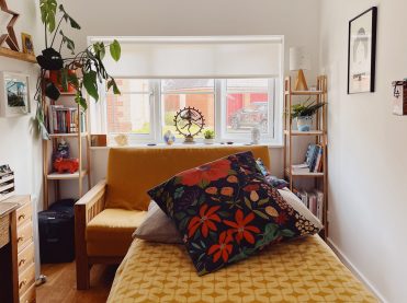 A white well lit room with a window at the end. In the foreground is a treatment couch with a yellow patterned blanket on it, a grey pillow and a navy cushion with lots of vibrant colours.